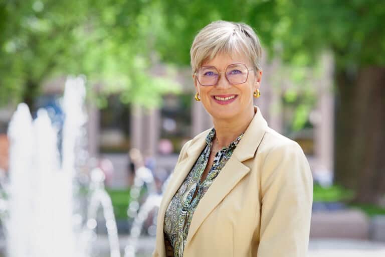 Moden kvinne smiler foran bypark med fontener, grønt treverk og oppdagende atmosfære, i en byromssetting.