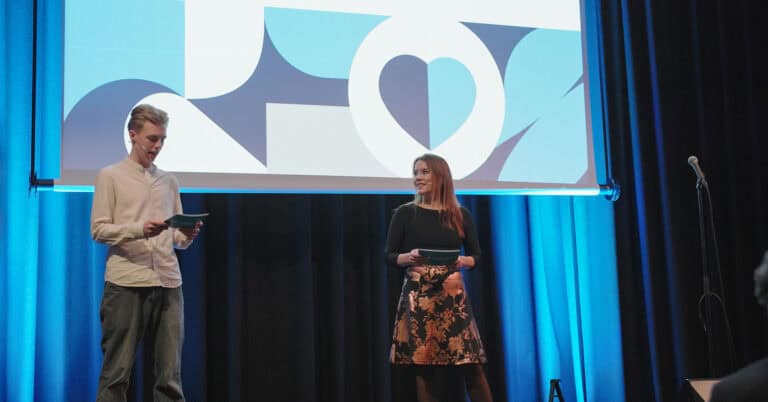 To personer på scen under presentasjon om byrom, med stor skjerm i bakgrunnen.