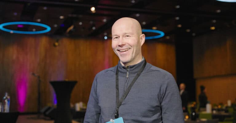 Mann smiler på konferanse i innendørs miljø.