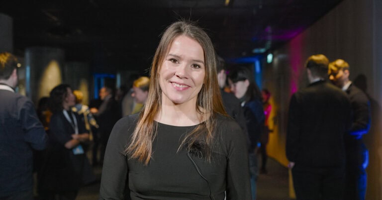 Hjerterom for byrom, kvinne smilende på sosialt arrangement, omgitt av mennesker i bakgrunnen.