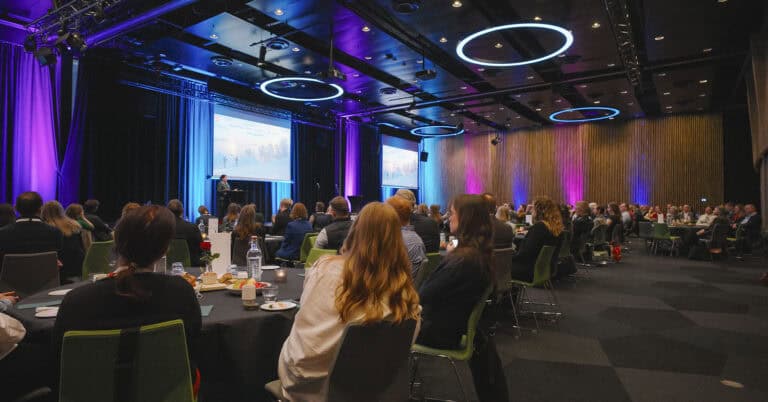 Deltakere på konferanse i stor, moderne sal med avansert lyd- og lysutstyr.