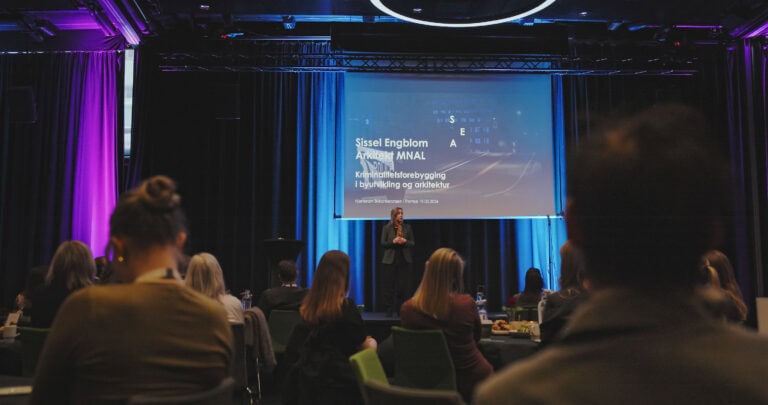 Person foran storskjerm under konferanse om byrom og innovasjon i et auditorium.