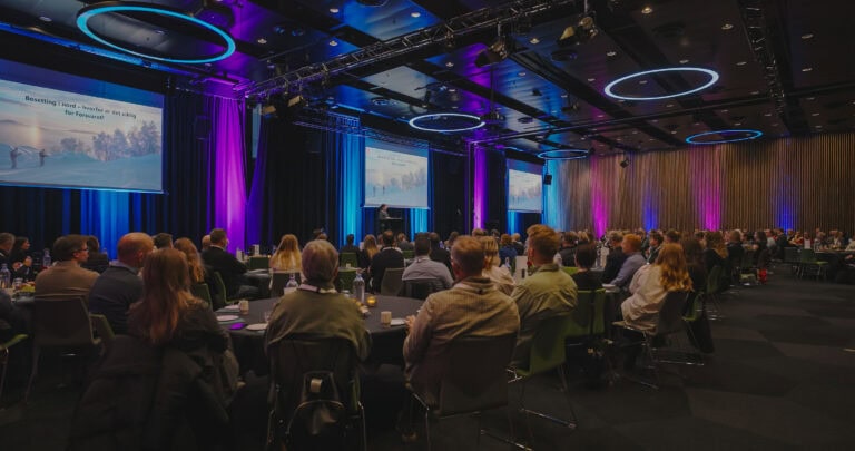 Publikum deltar på en konferanse i et moderne auditorium med store skjermer og fargerike lys.