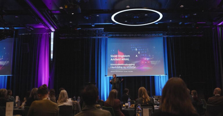 Publikum på konferanse i byrom med stor skjerm og scenelys.